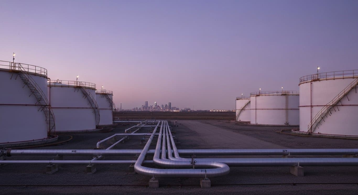 Midstream oil storage tanks and pipelines with a city skyline in the background under a purple sky