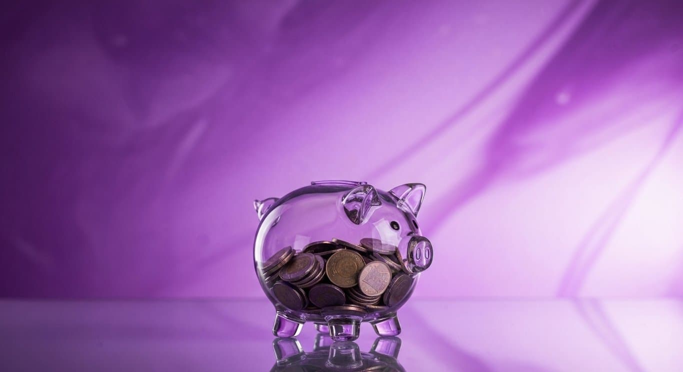 Glass piggy bank with coins on a reflective surface and a soft purple gradient background